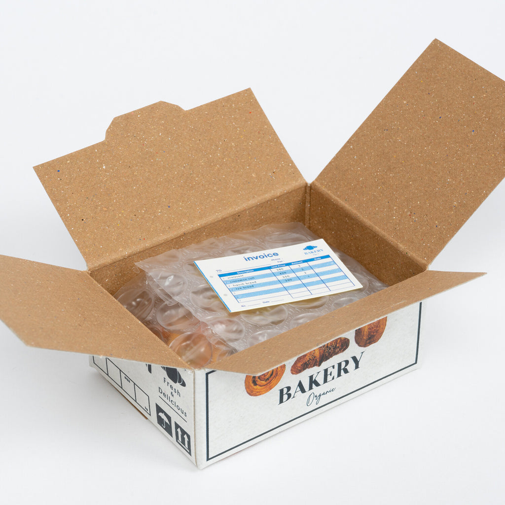Open cardboard box revealing protective packaging and an invoice, with bakery sticker designs visible on the side.