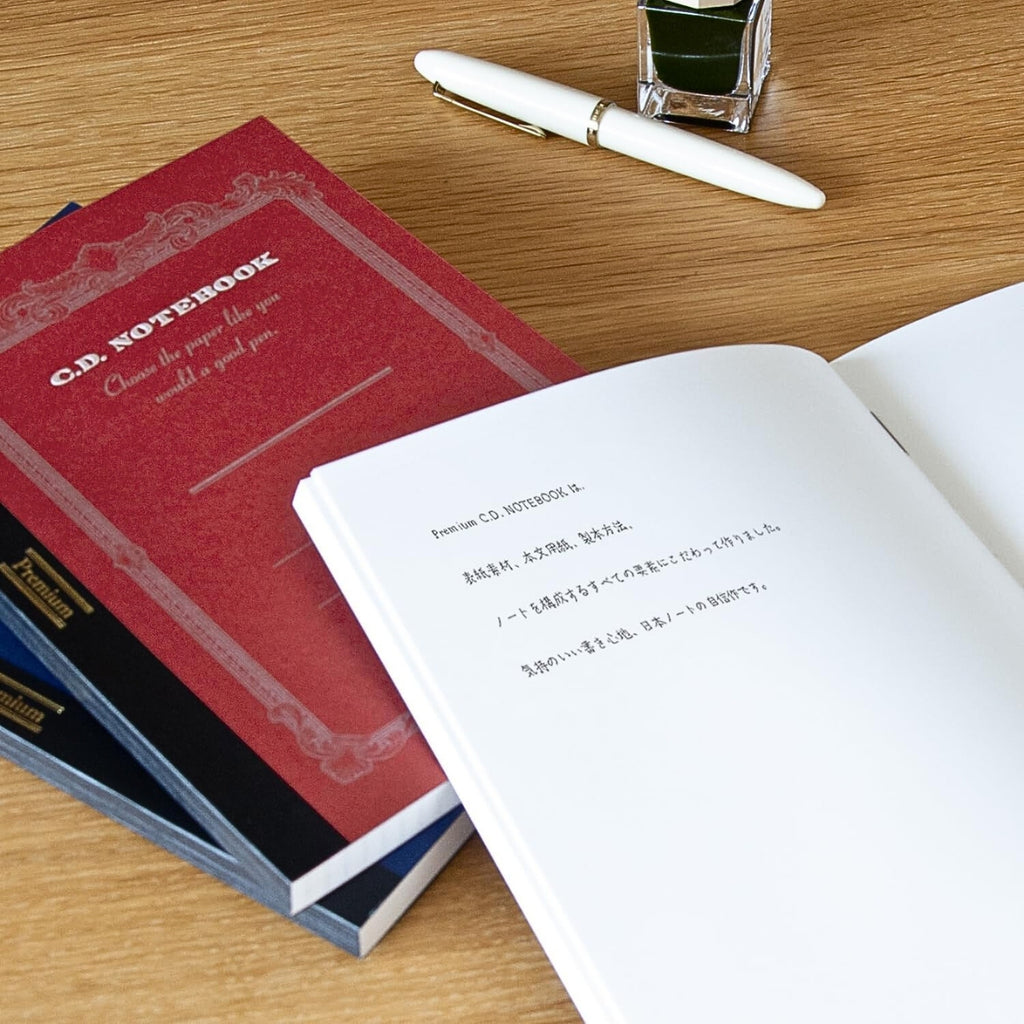 Apica Premium CD Notebooks in assorted colours shown open on a desk with writing tools and loose pages.