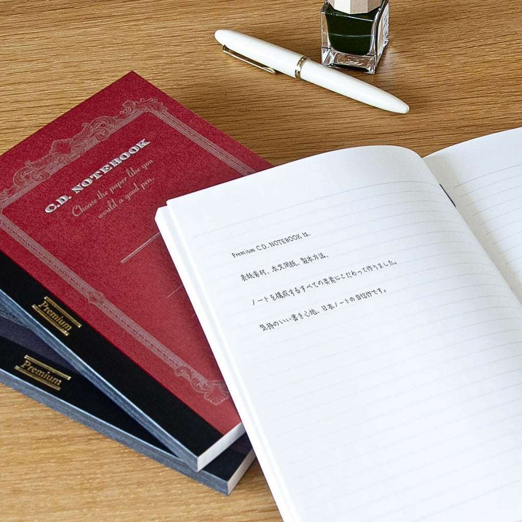 Apica Premium CD Notebooks in assorted colours shown open on a desk with pens and writing tools.