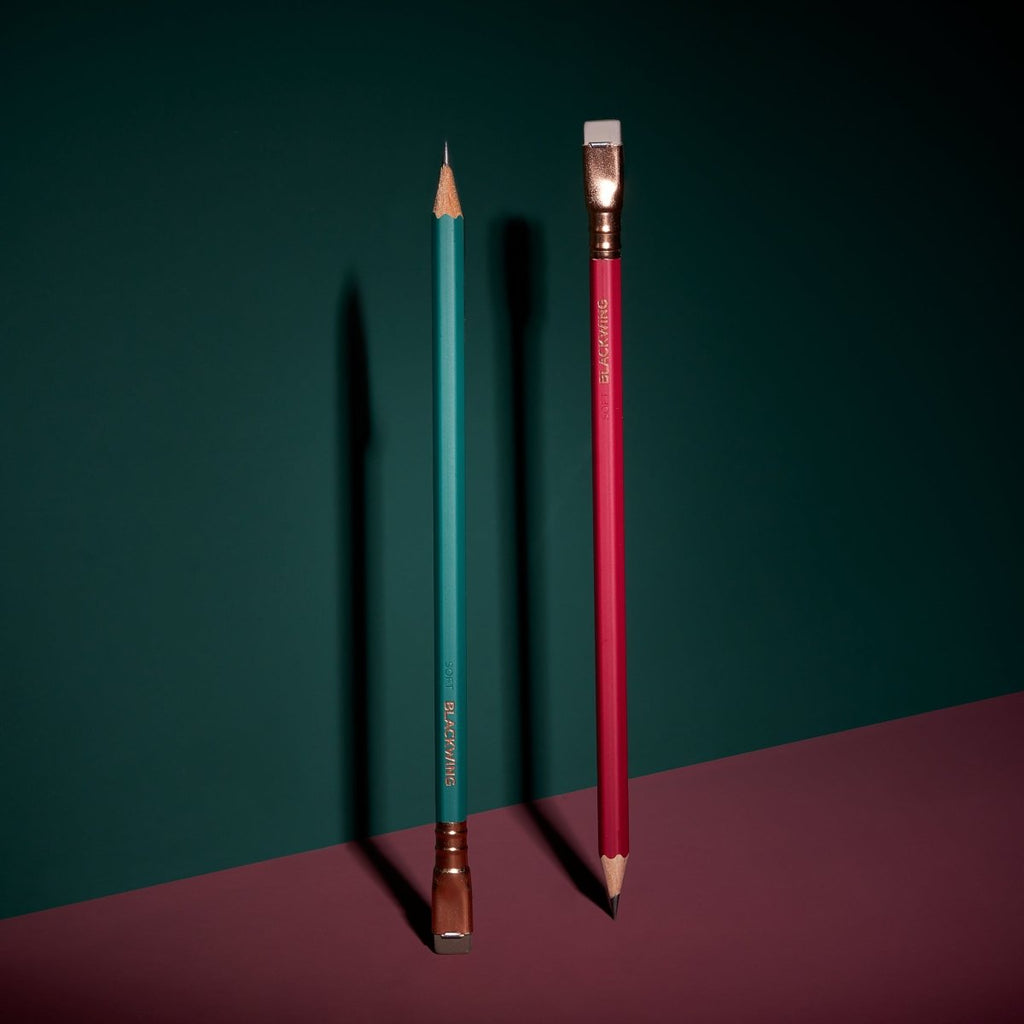 Two Blackwing Matte Red pencils standing upright on contrasting coloured surfaces.