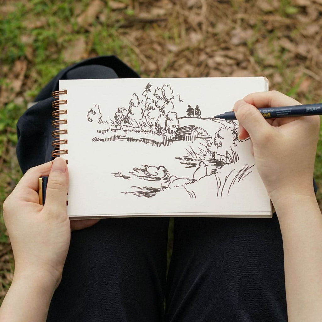 Person sketching a landscape scene in a Penco spiral-bound sketchbook while sitting outside.