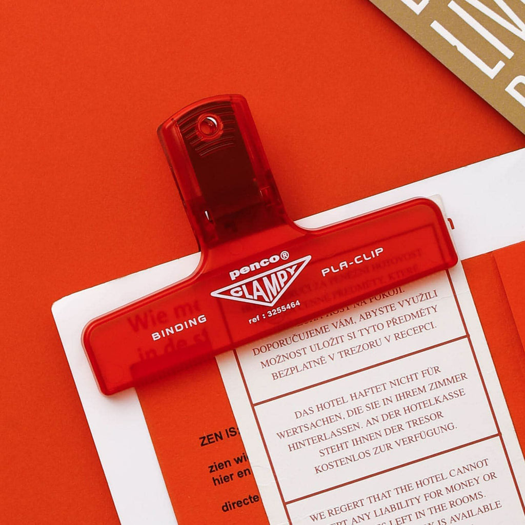 The Journal Shop - Red Hightide Penco Clampy Pla-Clip securing a printed sheet on an orange surface.