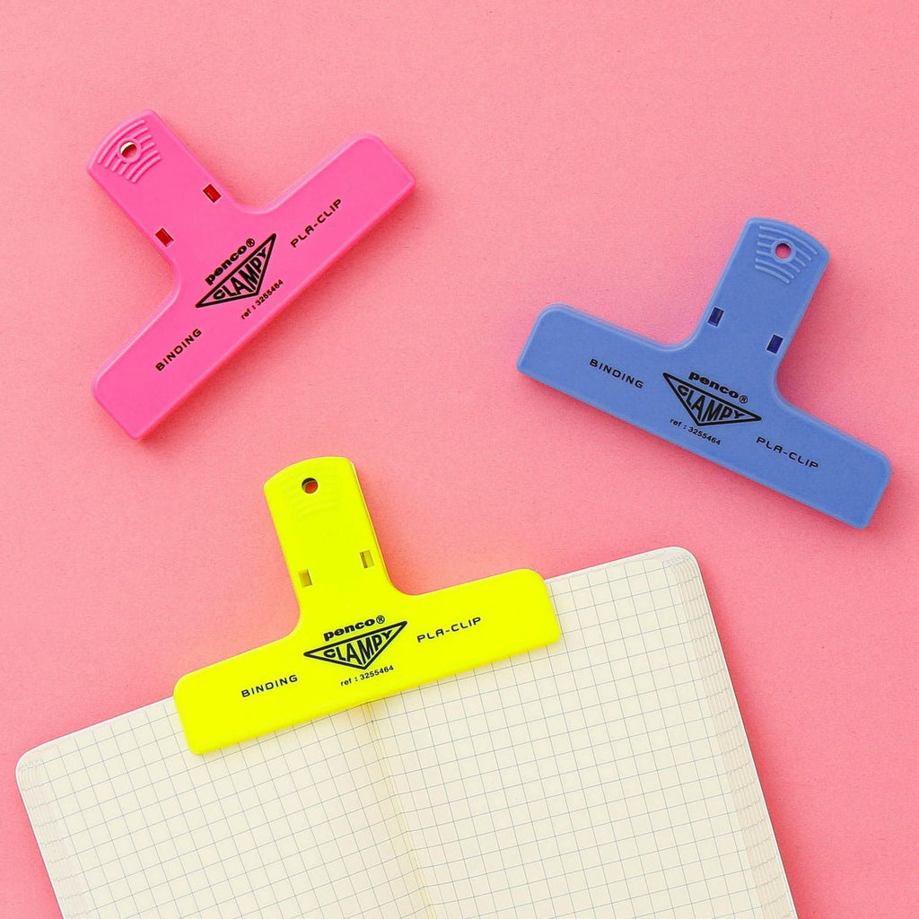 The Journal Shop - Trio of Hightide Penco Clampy Pla-Clips in pink, neon yellow, and blue attached to lined notepaper on a pink desk.