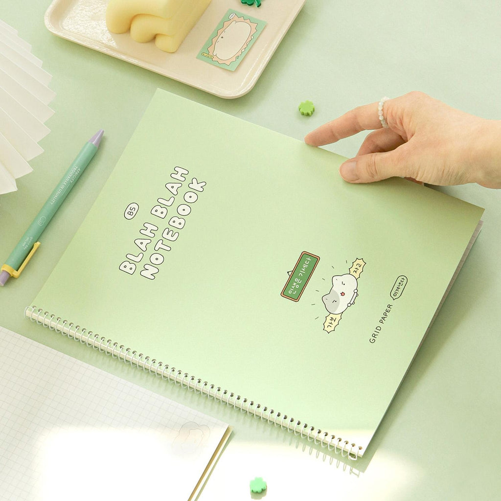 The Journal Shop - Iconic Blah Blah B5 Spiral Grid Notebook green cover displayed on desk being lifted by hand.
