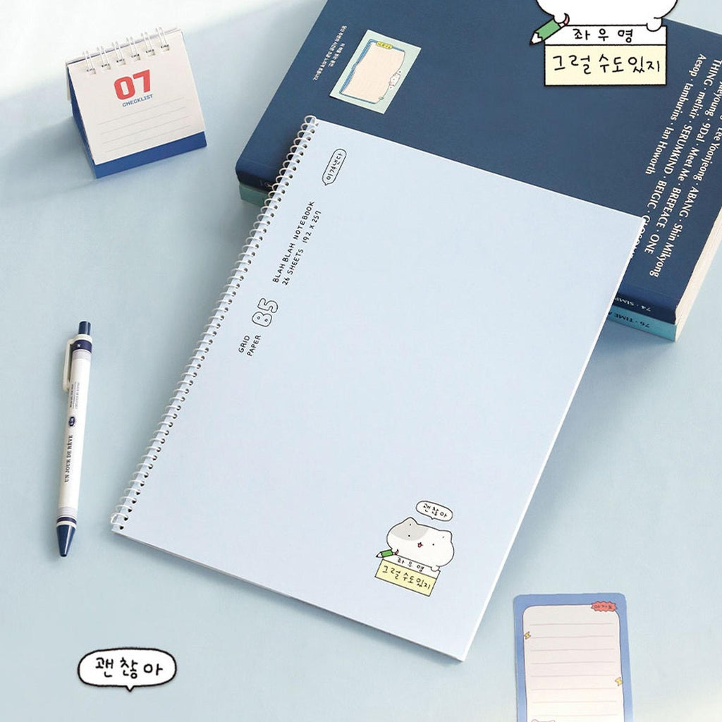 The Journal Shop - Iconic Blah Blah B5 Spiral Grid Notebook blue cover open beside pen and paper clips on white surface.