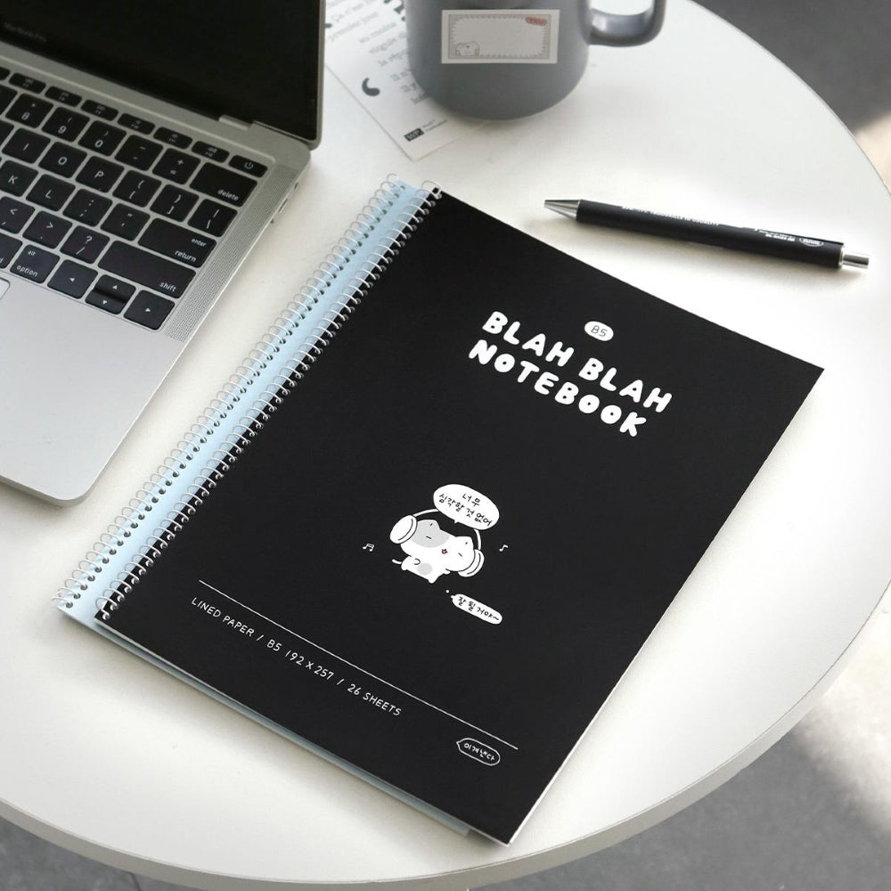 The Journal Shop - Iconic Blah Blah B5 Spiral Lined Notebook black cover with white illustration placed beside laptop and coffee cup.