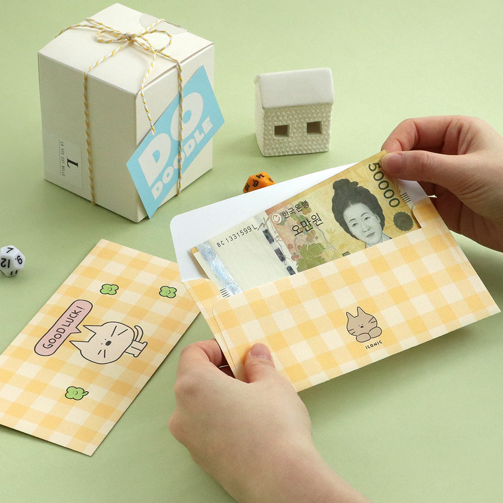 The Journal Shop - Hands placing Korean won banknotes into a yellow Iconic Doodle Envelope featuring a cute cartoon cat and “Good Luck!” message, beside a wrapped gift box and small decorative items on a light green surface.