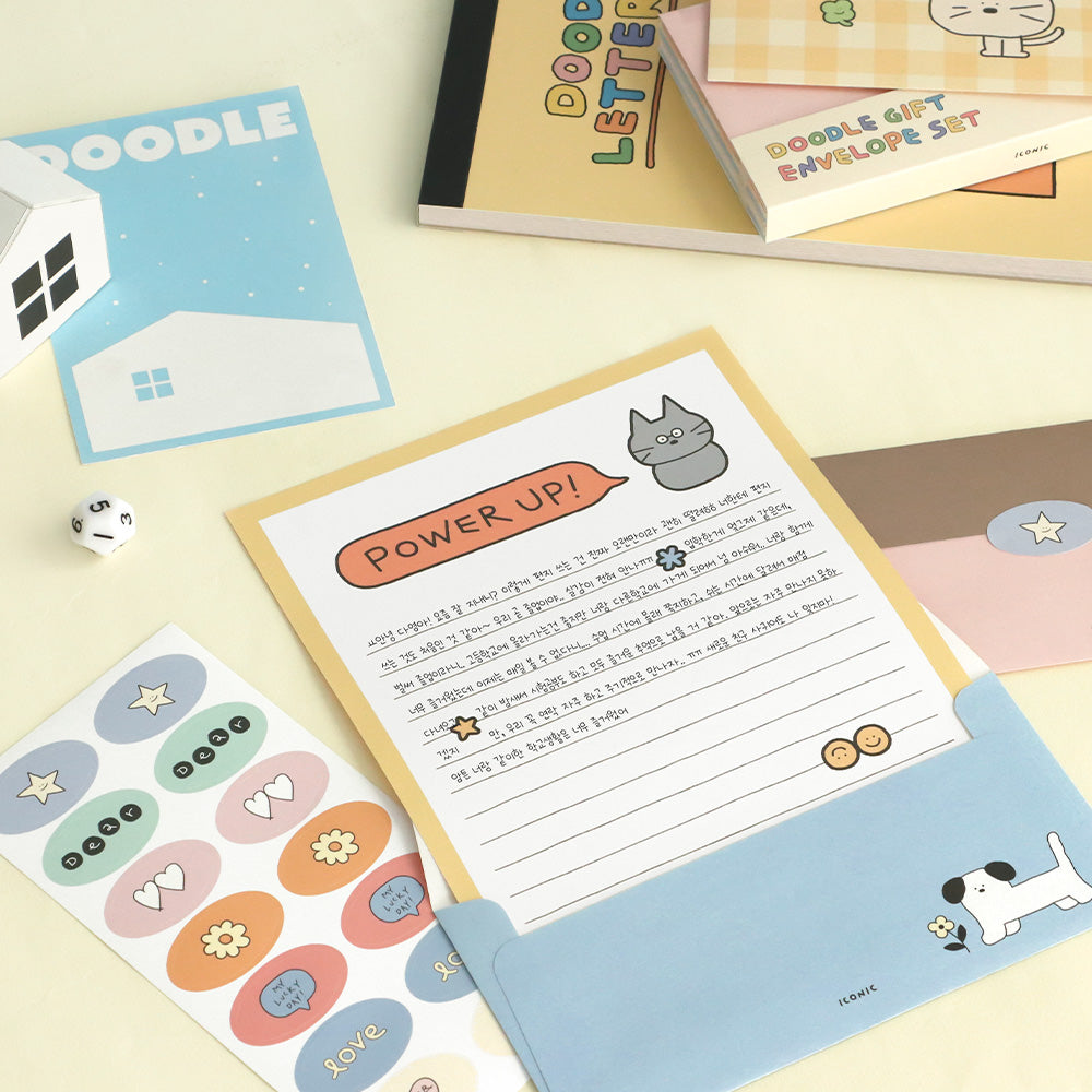 The Journal Shop - Flat lay of Iconic Doodle stationery featuring a “Power Up!” illustrated letter sheet in a blue envelope with a cartoon dog, surrounded by sticker sheets, notebooks, and envelopes from the Doodle Gift collection on a light yellow surface.