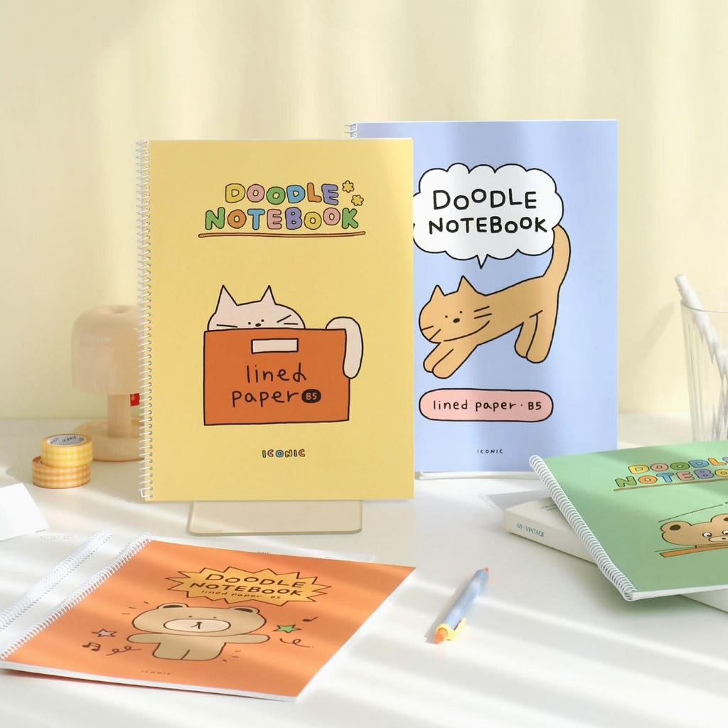 The Journal Shop - Iconic Doodle B5 Spiral Lined Notebooks in various colours with illustrated covers stacked and displayed on table.