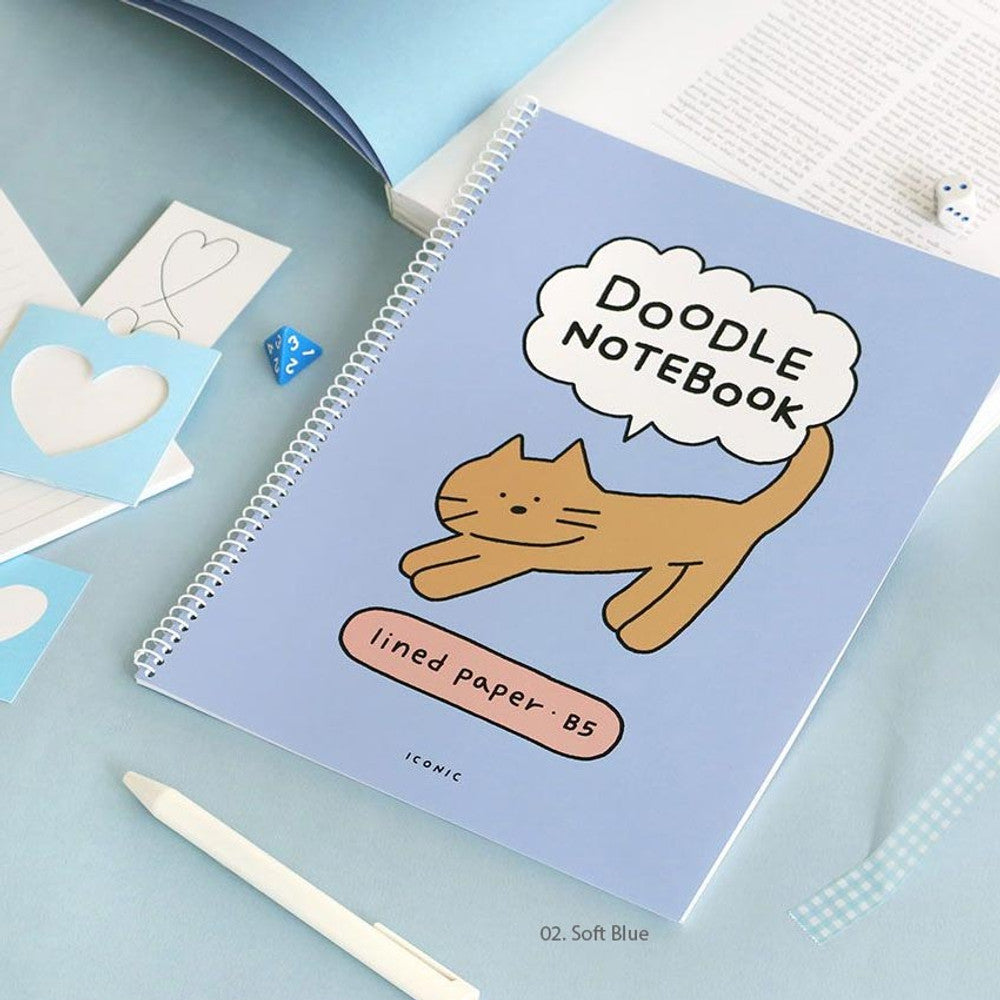 The Journal Shop - Iconic Doodle B5 Spiral Lined Notebook in blue cover open on pastel background with writing utensils.