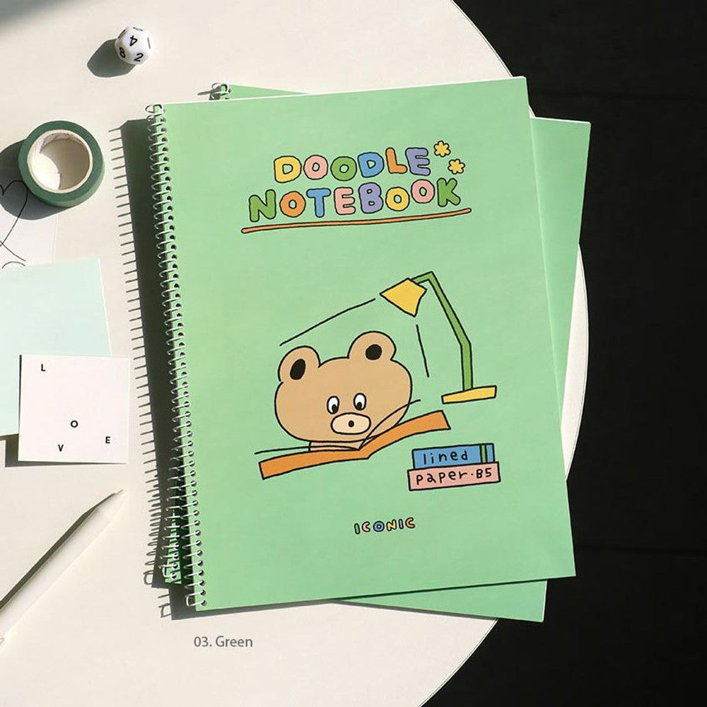 The Journal Shop - Iconic Doodle B5 Spiral Lined Notebook in green cover with illustrated character, lay flat on desk with stationery tape.