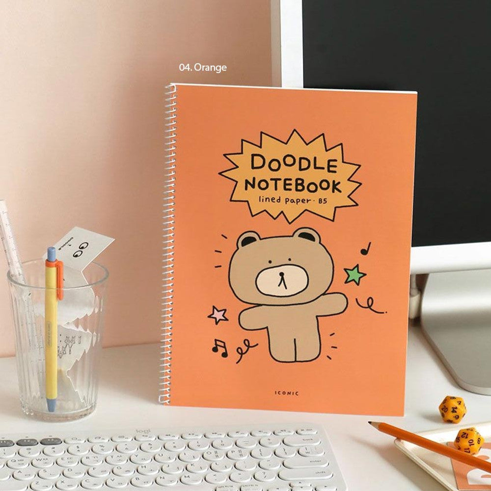 The Journal Shop - Iconic Doodle B5 Spiral Lined Notebook in orange cover with illustrated character, shown next to laptop on desk.
