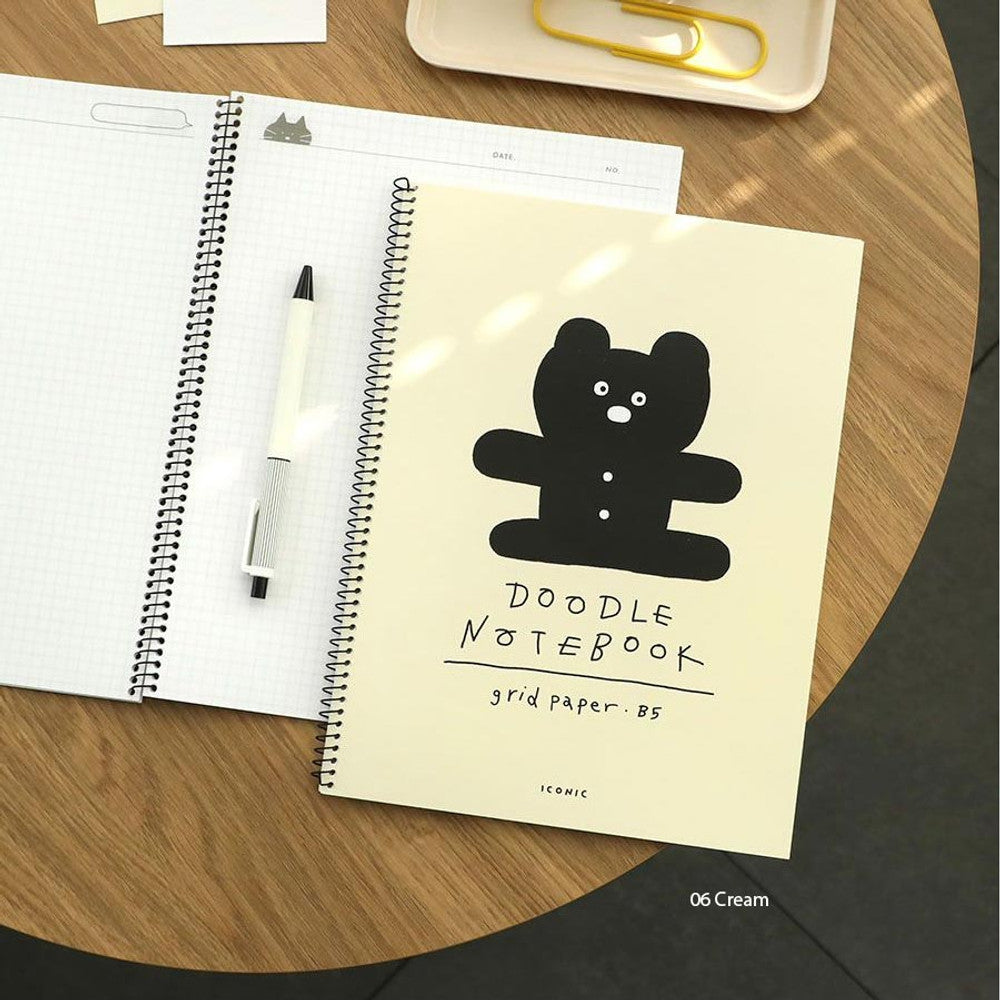 The Journal Shop - Iconic Doodle Spiral B5 Grid Notebook in beige and black covers displayed on wooden desk with pen.