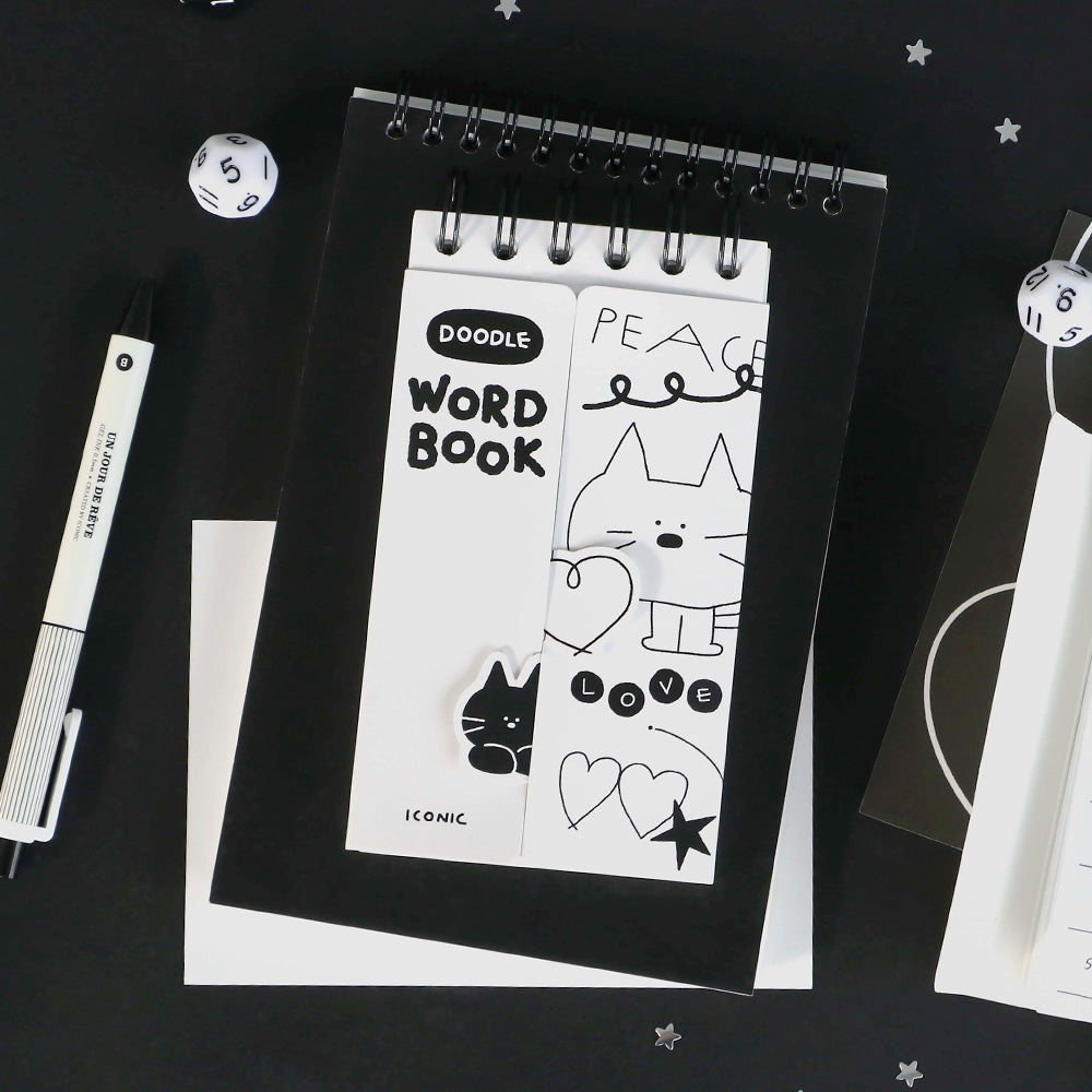 The Journal Shop - A white Iconic Doodle Word Pocket Size Notebook with playful doodle-style characters, shown on a dark workspace beside a pen.