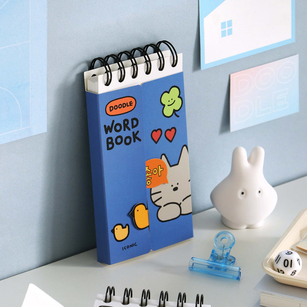 The Journal Shop - A blue Iconic Doodle Word Pocket Size Notebook standing upright on a tidy desk beside stationery and a small white rabbit figurine.