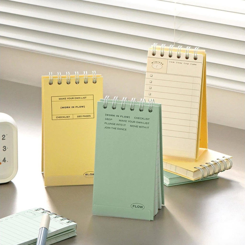 The Journal Shop - Two Iconic Flow Checklists in yellow and mint green standing on a bright desk by a window, with light streaming through the blinds.