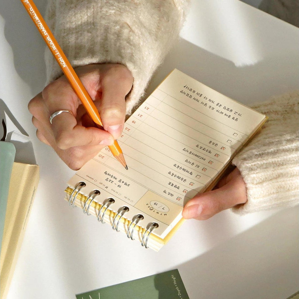 The Journal Shop - A person writing in the yellow Iconic Flow Checklist while resting it on a desk, with a pencil in hand.