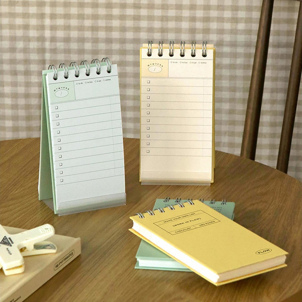 The Journal Shop - A pair of Iconic Flow Checklists in mint green and yellow placed on a wooden table with paper clip and notebooks.