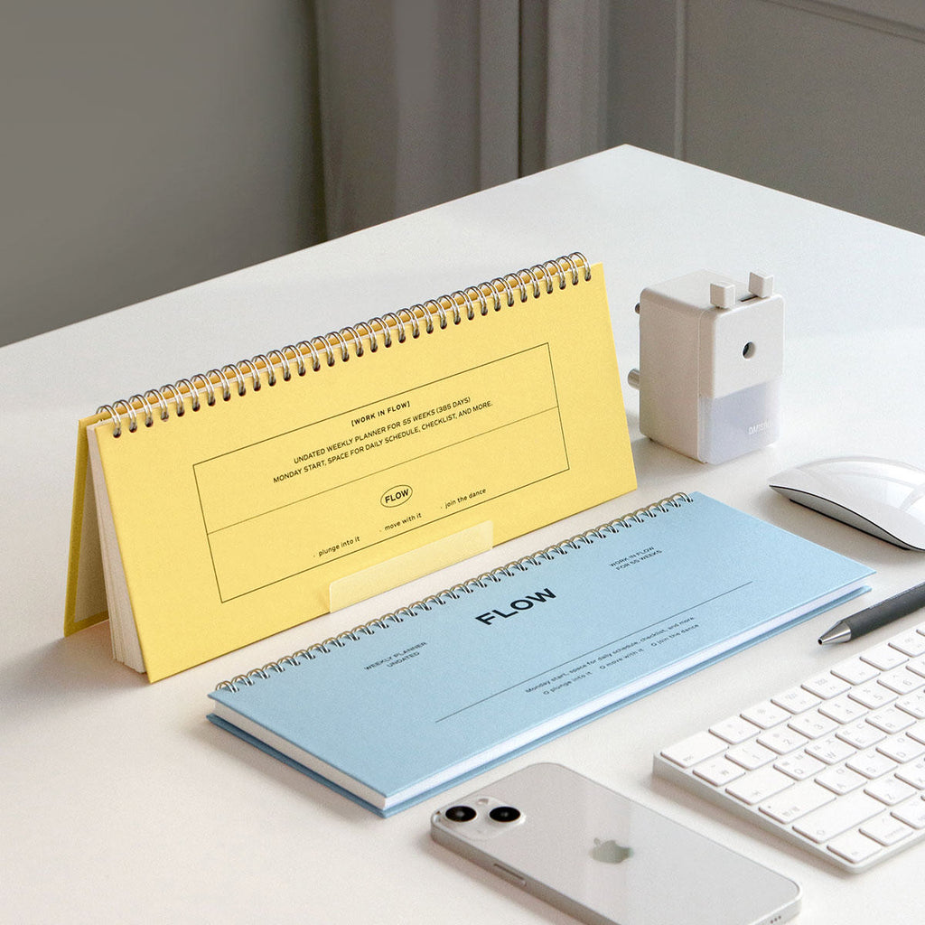The Journal Shop - Yellow and blue Iconic Flow Weekly Planners displayed on a white desk beside a keyboard, phone, and pen.
