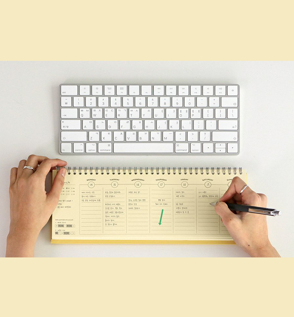 The Journal Shop - Hand holding a pen over the butter yellow Iconic Flow Weekly Planner next to a white keyboard.
