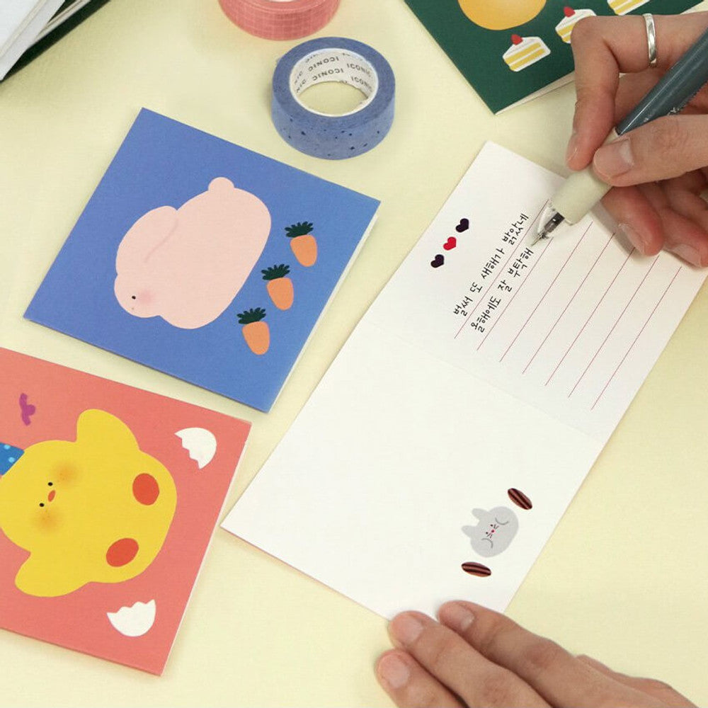 The Journal Shop - Person writing on Iconic Happy Mini Card with illustrated animal design beside open box and stationery.