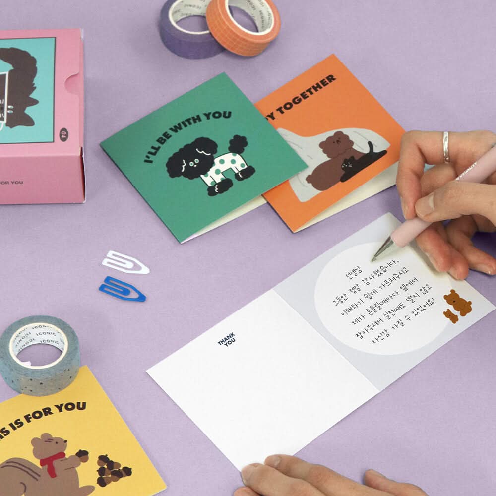 The Journal Shop - Person writing on Iconic Mini Friends Card featuring dogs design beside stationery accessories.