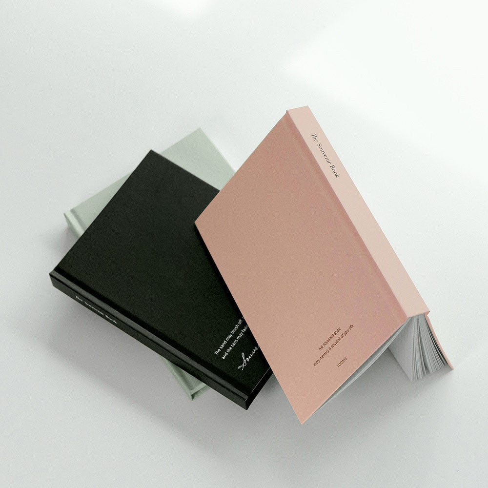 The Journal Shop - A pastel pink Iconic Souvenir B6 Grid Notebook resting open on a desk with a black notebook placed beside it.