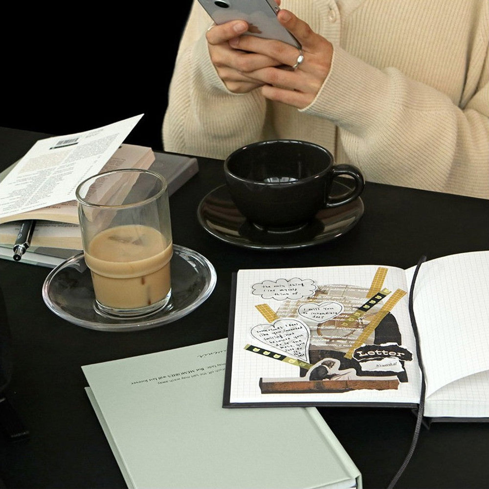The Journal Shop - A person writing in an Iconic Souvenir B6 Grid Notebook while enjoying a drink at a café table.