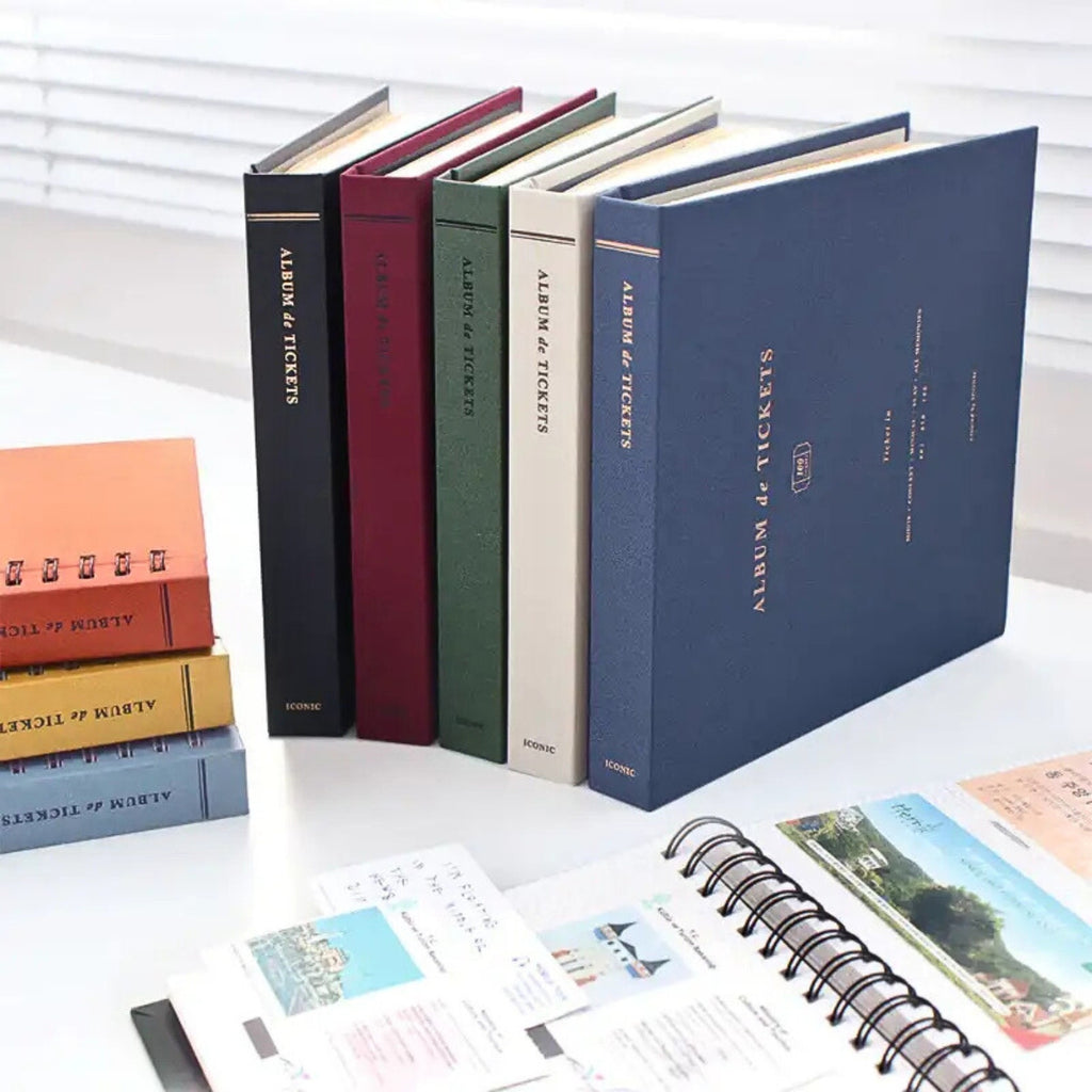 Stack of Iconic Ticket Scrapbook Albums in assorted colours standing upright on a desk with stationery items.