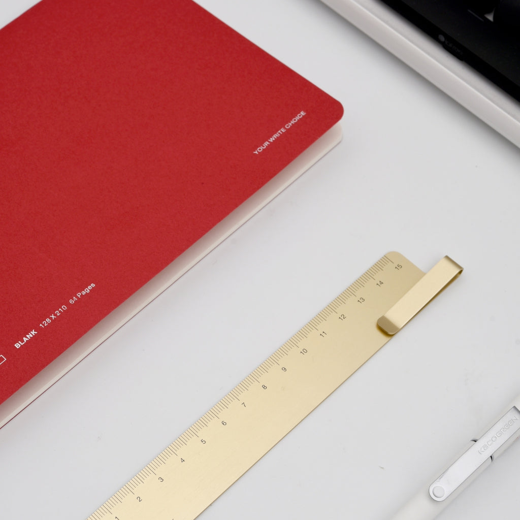 Gold metal ruler with clip alongside a red notebook and white pen on a clean desk.