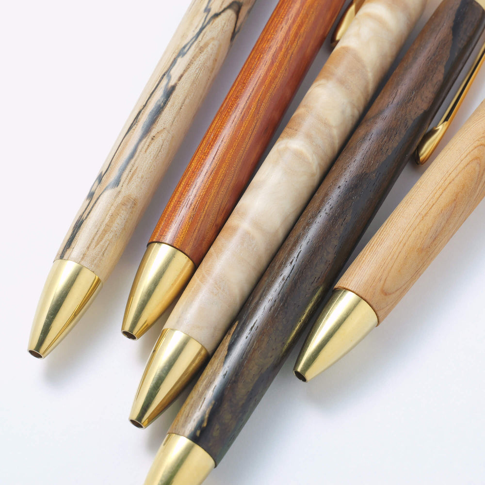 The Journal Shop - Collection of Kakimori Handcrafted Ballpoint Pens in assorted wood finishes with brass hardware, arranged in a lay flat display.