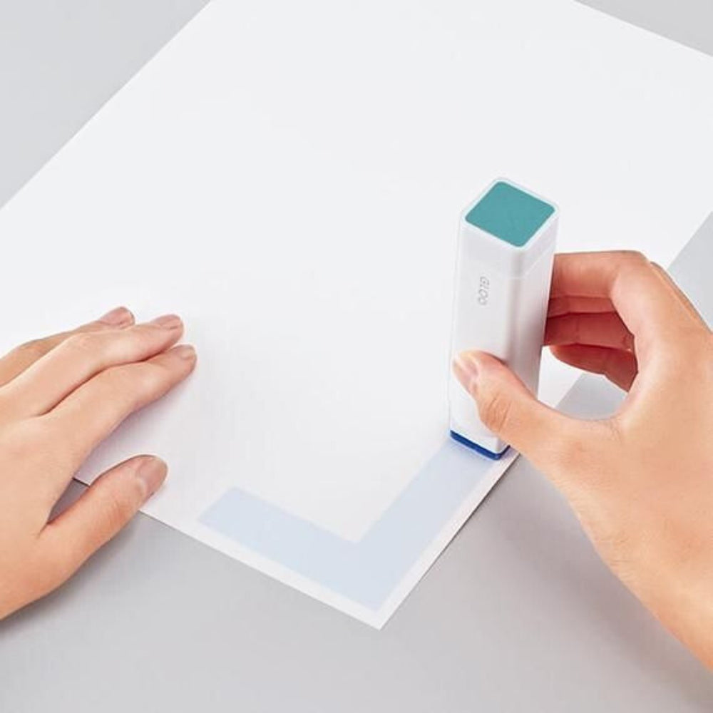 Hand using KOKUYO GLOO glue stick to apply adhesive to the corner of a white sheet.