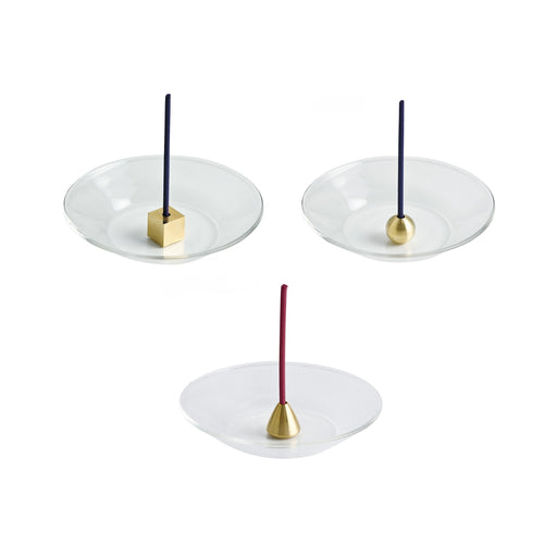 Brass cube incense holder placed on a clear glass plate with a lit incense stick standing upright.