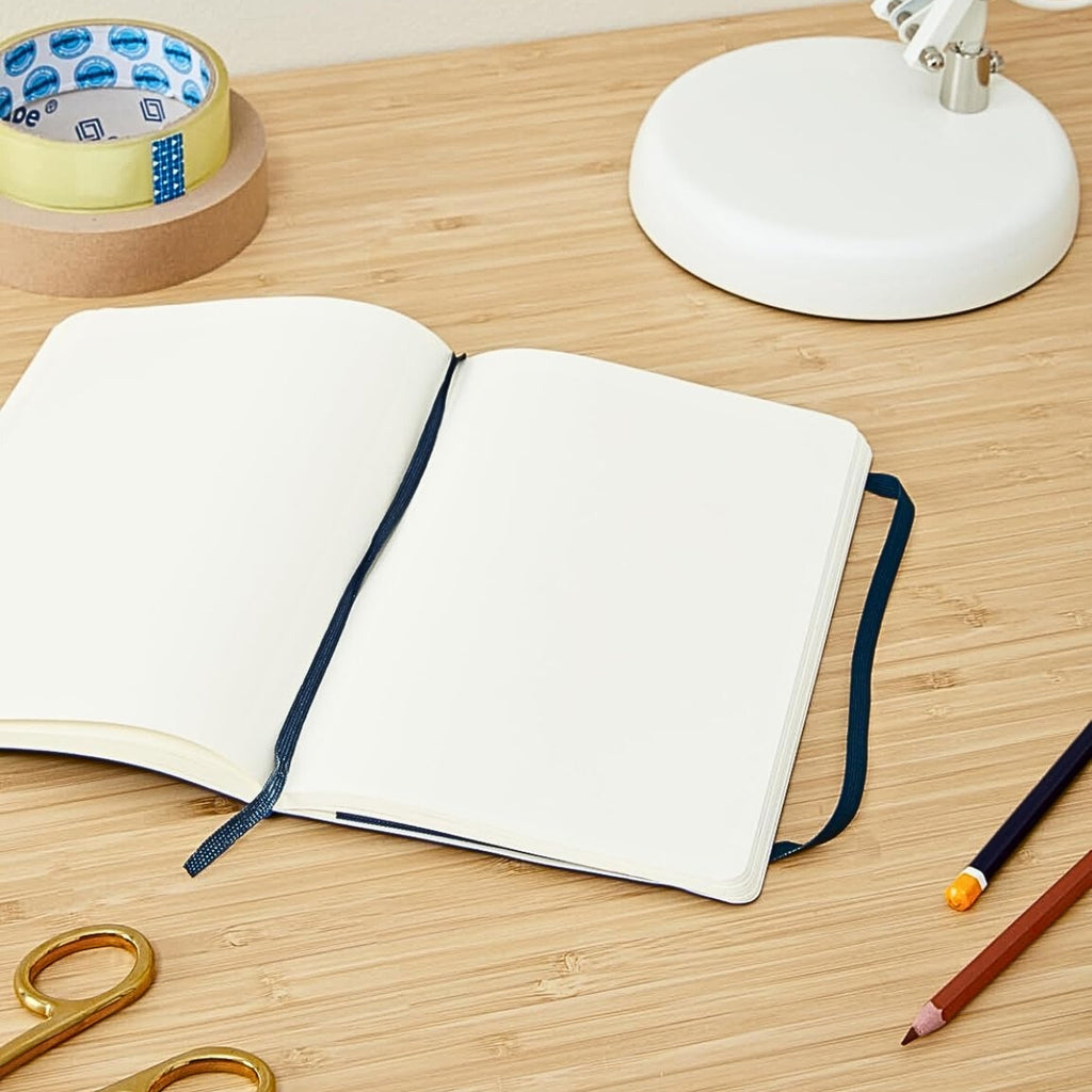 Open Moleskine notebook on a wooden desk with stationery and lamp nearby.