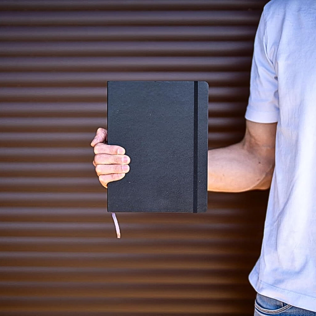 Person holding a Moleskine Extra Large Black Cahier Dotted notebook outdoors against metal wall.