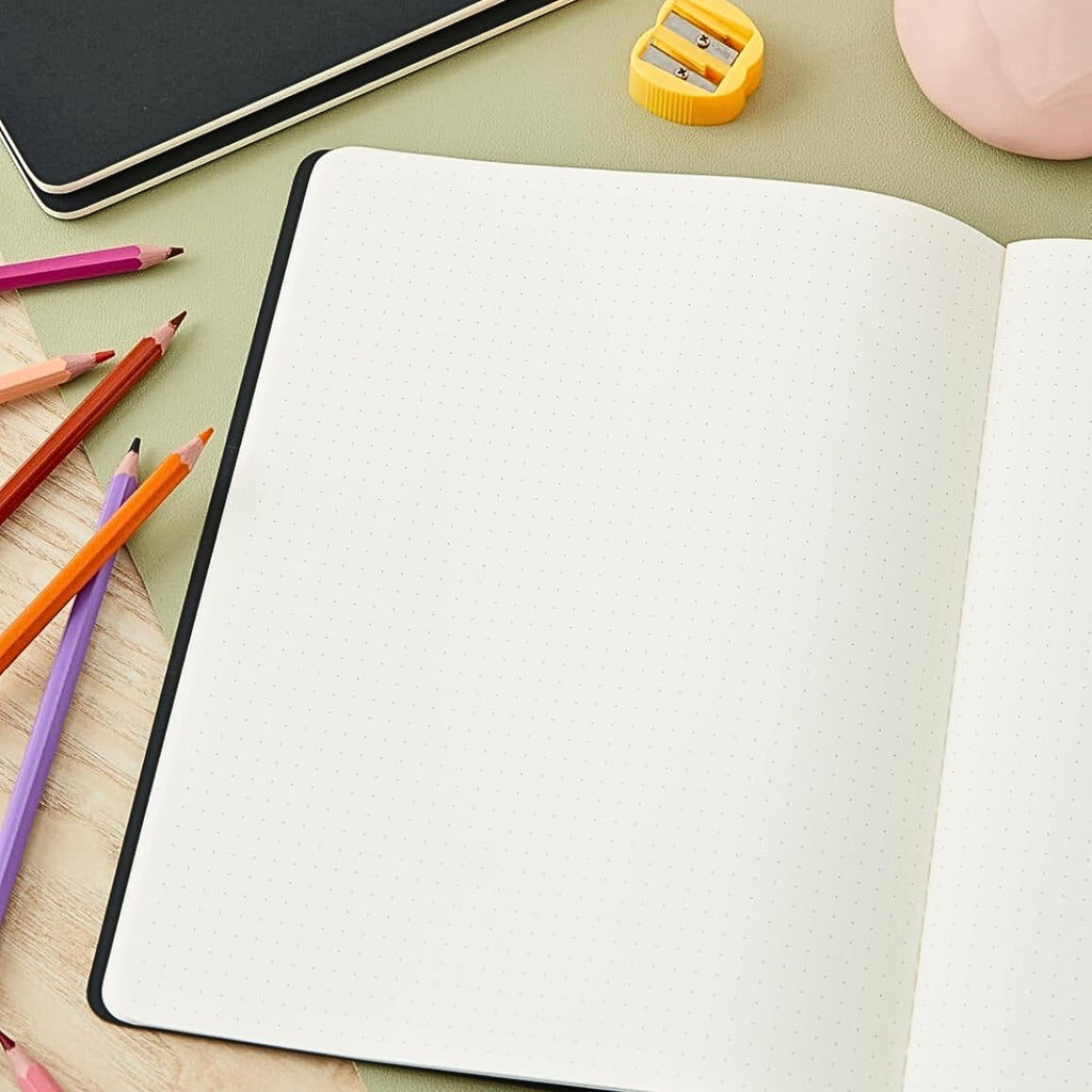 Open Moleskine Extra Large Black Cahier Dotted notebook beside pencils on a desk.