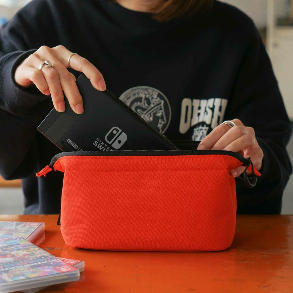 A person is holding a red soft gadget pouch with a Nintendo Switch inside it, which is placed on top of a table with a few books scattered around.