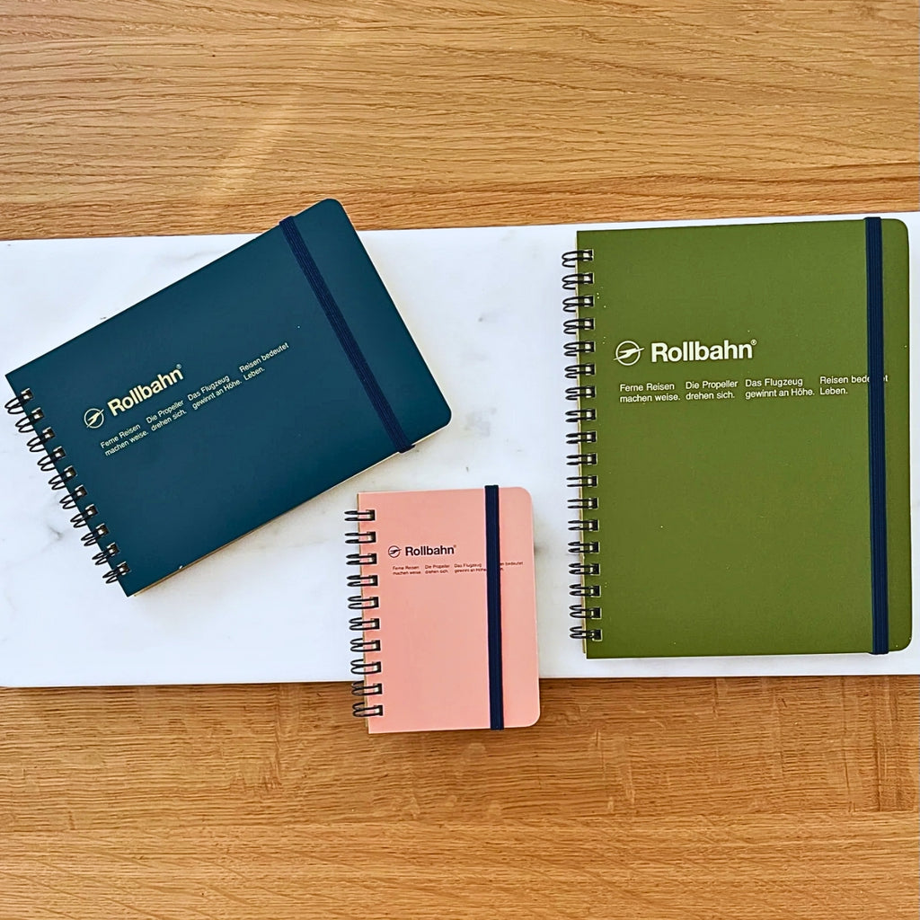 A selection of three Rollbahn spiral notebooks in various sizes and colours, arranged on a wooden and marble surface.